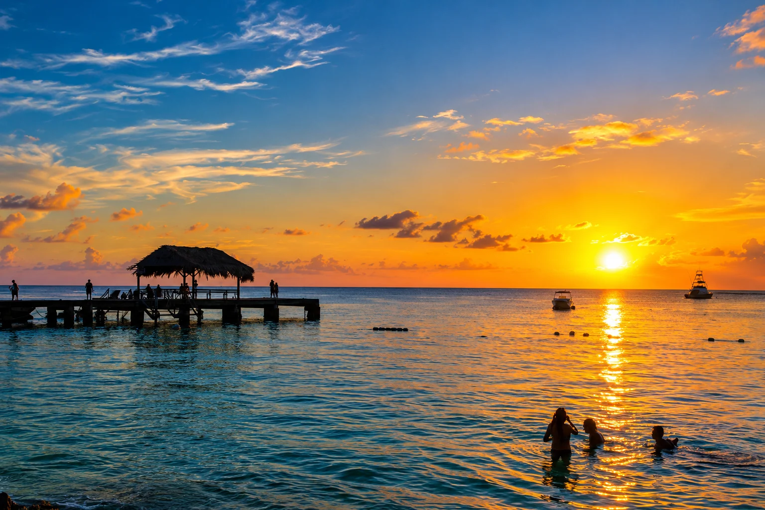 Sunset over a bay with a thatched-roof jetty in silhouette and swimmers in the water