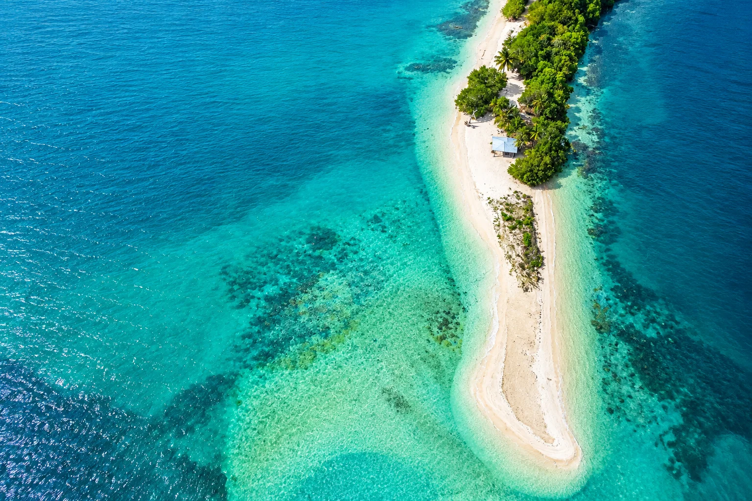 Aerial of a narrow wooded sand spit running into turquoise reef shallows