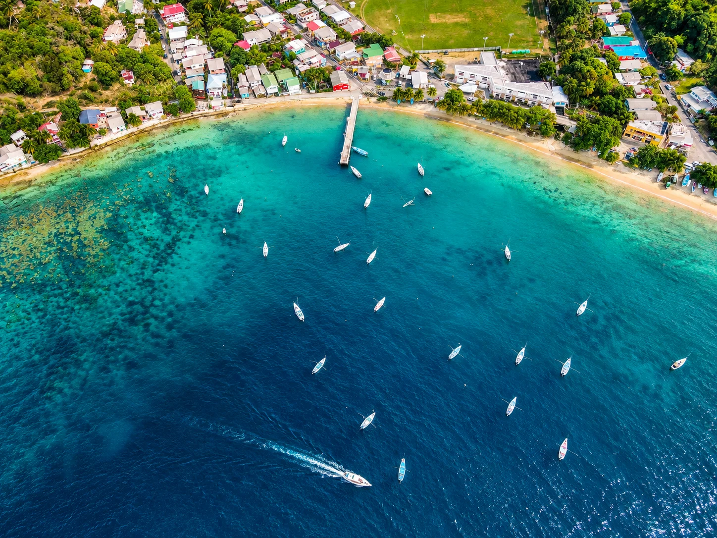 Top-down aerial of a village bay with jetty and many small fishing boats moored over turquoise water